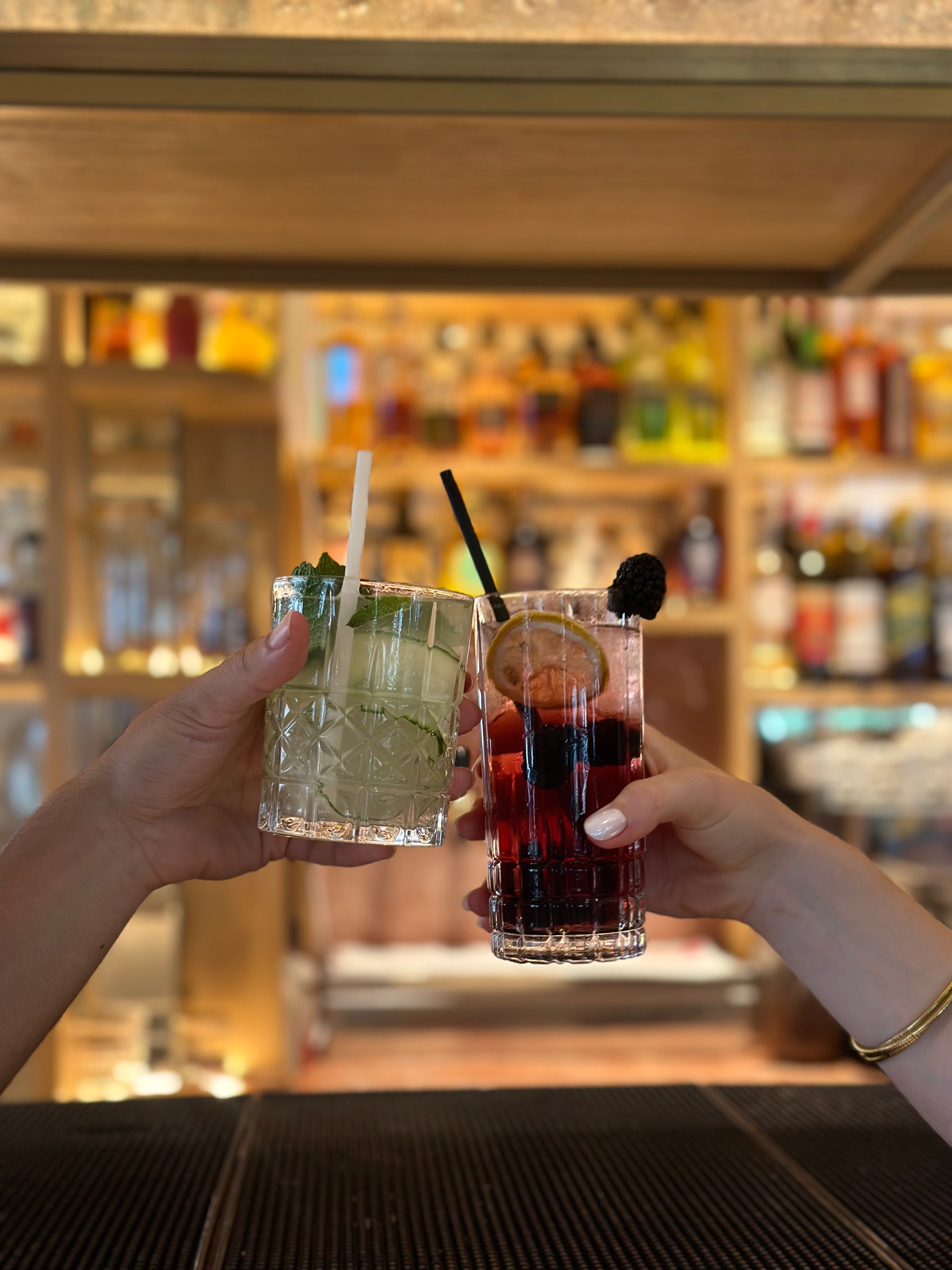 deux mains qui trinquent devant un bar professionnel en arrière plan avec  deux pailles Strws-berry différentes et deux cocktails différents : un short drink et un long drink. Paille noir roche et paille blanc givré